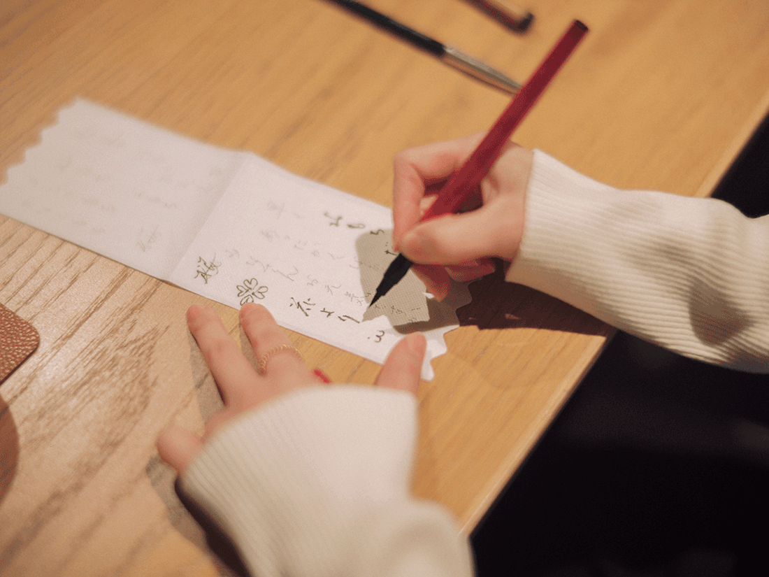 木のテーブルで筆文字を書く手元のアップ。丁寧な創作風景