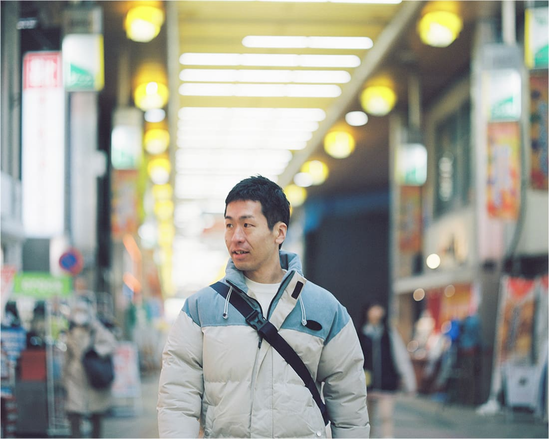 商店街を歩く男性。明るい照明が続く通りの中、穏やかに微笑んでいる。