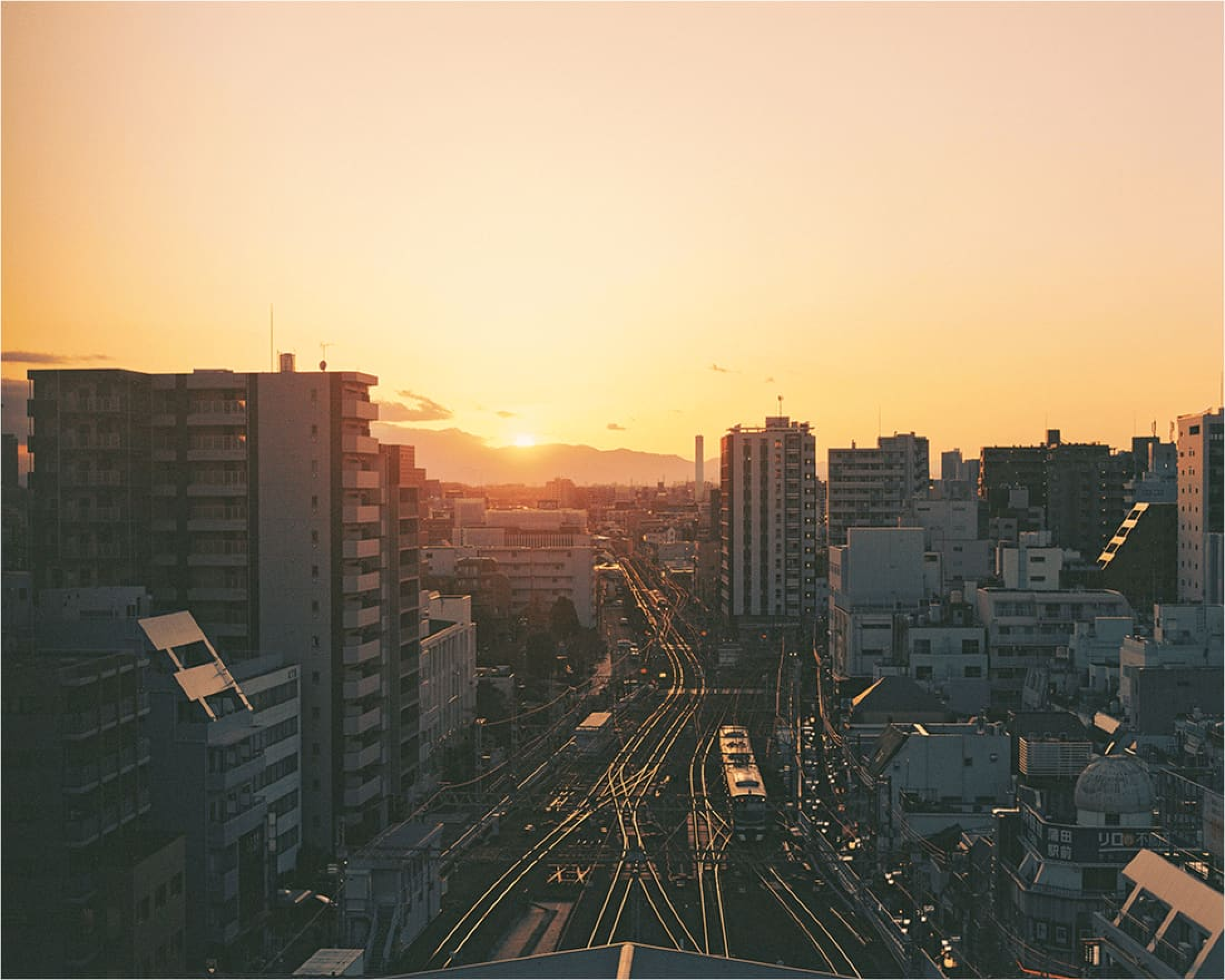 夕暮れ時の蒲田の街並みを高所から望む。遠くに山が見え、鉄道の線路が夕日に照らされて輝いている。