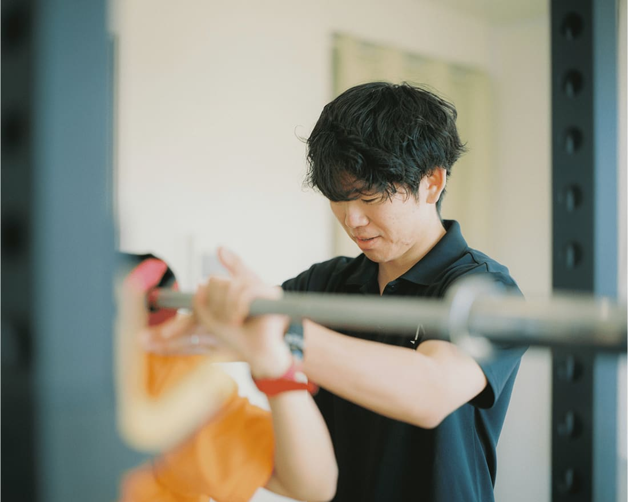 バーベルを支える男性。集中した表情で、クライアントのトレーニングを見守っている。